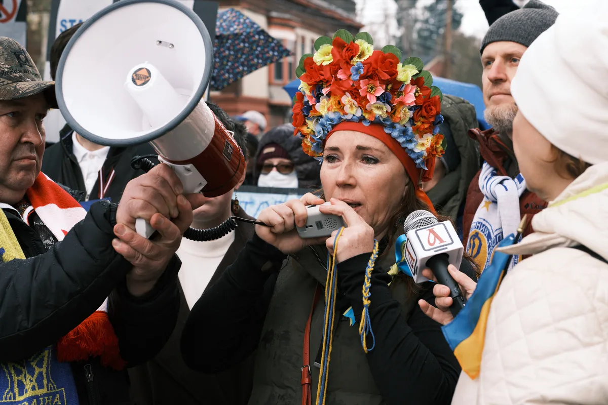 A woman crowned with an elaborate headdress of vibrant artificial flowers—red roses mingling with blue and yellow blooms—speaks into multiple microphones thrust toward her by surrounding media and protesters. Her weathered face reflects determination as she addresses the crowd outside Portland's Revolution Hall, wrapped in dark winter clothing against the gray February chill. The scene pulses with urgent energy as megaphones and recording devices create a constellation of amplification around her impassioned words, while Ukrainian flag colors echo symbolically in her floral crown.