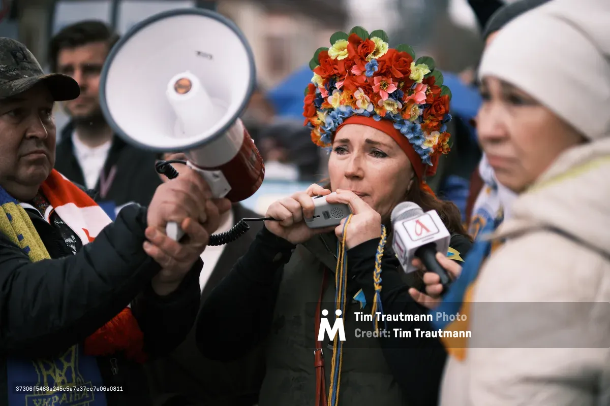 A woman crowned with an elaborate headdress of crimson roses and azure blooms speaks passionately into news microphones outside Portland's Revolution Hall, her voice amplified by a white bullhorn held aloft by a supporter. The scene captures the urgent atmosphere of an anti-war demonstration, with protesters bundled in winter coats against the Oregon chill, their faces etched with determination as media microphones cluster around the floral-adorned activist. The soft, overcast lighting creates an intimate yet charged moment of civic engagement in the days before international conflict would escalate.