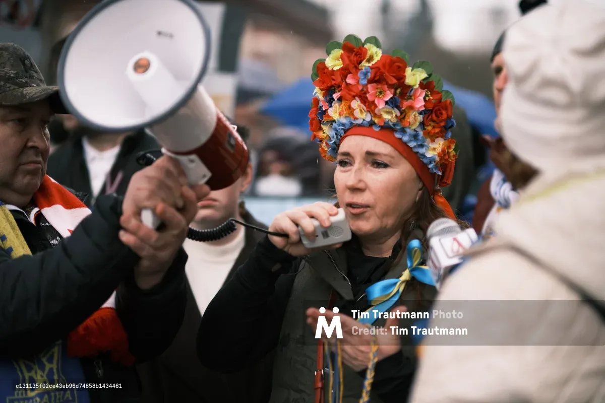 A woman crowned with an elaborate headdress of vibrant artificial flowers—crimson roses, azure blooms, and golden petals—speaks passionately into microphones as protesters gather outside Portland's Revolution Hall. The ornate floral crown creates a striking juxtaposition against the serious nature of her anti-war message, while megaphones and recording devices thrust toward her capture the urgency of the moment. The scene pulses with the electric tension of impending conflict, documented just days before Russian forces would cross into Ukraine.