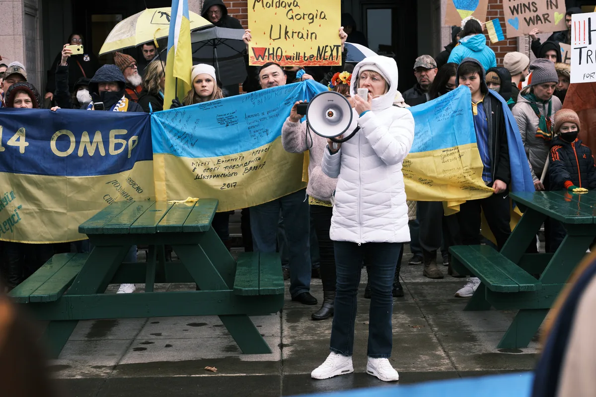 A determined protester in a white puffy jacket amplifies her message through a megaphone, standing before a weathered Ukrainian flag inscribed with signatures and messages of support. The scene unfolds on the rain-dampened concrete plaza outside Portland's Revolution Hall, where dozens of bundled demonstrators have gathered in the gray winter light. Handmade signs warning "WHO will be NEXT" pierce through the crowd, creating a prophetic tableau of solidarity captured just days before Russia's invasion began.
