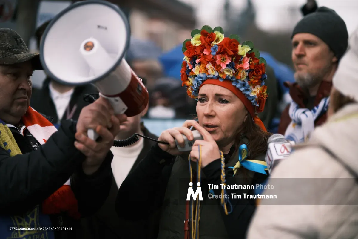 A woman adorned with a vibrant floral headdress—crimson roses and azure blooms echoing Ukrainian colors—speaks into a microphone at an anti-war demonstration outside Portland's Revolution Hall. Her passionate expression cuts through the crowd of bundled protesters, while a megaphone hovers nearby, amplifying voices of dissent in the cold Oregon air. The intimate gathering pulses with urgency, captured just days before Russian forces would cross into Ukraine.