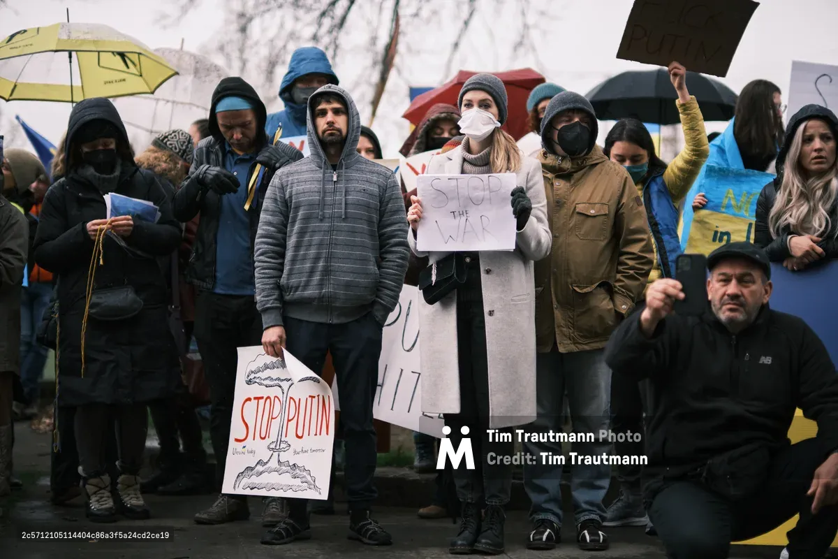 Protesters gather beneath umbrellas and winter coats outside Portland's Revolution Hall, their signs calling for an end to war as bare branches frame the scene above. A woman in a light coat holds a handwritten "STOP THE WAR" placard while masked demonstrators clutch Ukrainian flags and anti-Putin messaging, their collective breath visible in the cold February air. The overcast sky and wet pavement reflect the somber urgency of their cause, captured just days before Russia's invasion of Ukraine would transform their peaceful plea into prophetic witness.