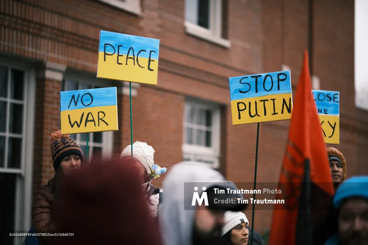 Bundled against Portland's winter chill, protesters gather outside Revolution Hall's historic brick facade, their handmade signs bearing urgent messages of peace. The Ukrainian flag colors of blue and yellow dominate the placards reading 'PEACE,' 'NO WAR,' and 'STOP PUTIN,' while an orange banner cuts through the crowd like a warning flame. The shallow depth of field isolates the passionate faces and bold typography against the weathered brick of the Central Eastside landmark, capturing a moment of civic urgency in the days before conflict erupted.