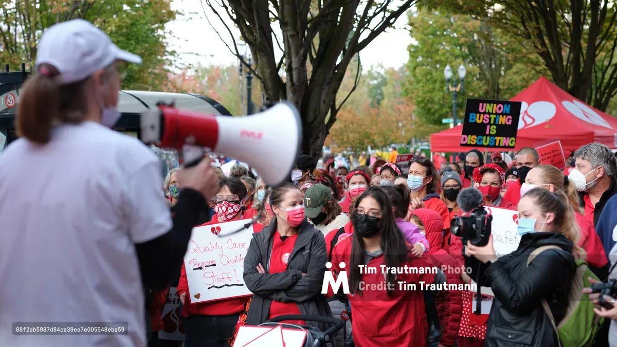 A sea of masked healthcare workers in crimson scrubs and winter jackets gather beneath autumn trees in Portland's Lloyd District, their unified stance punctuated by handmade signs demanding quality care and fair staffing. A speaker with a red and white megaphone commands attention from the foreground, while the crowd extends toward a vibrant tent displaying union solidarity messages. The overcast sky and golden fall foliage create a dramatic backdrop for this moment of collective action outside Kaiser Permanente's tower.