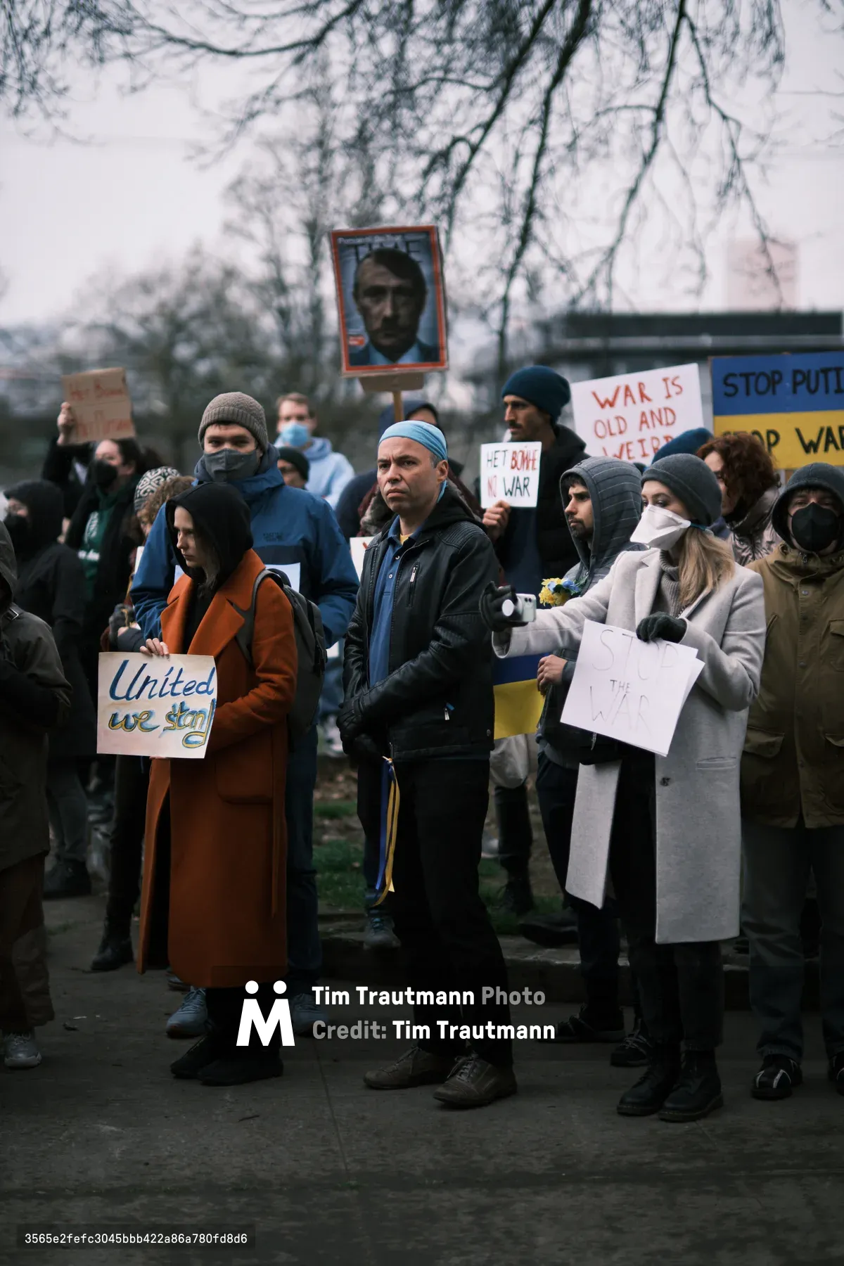 Bundled against Portland's winter chill, protesters gather beneath skeletal tree branches outside Revolution Hall in the city's Central Eastside district. A man in a blue surgical cap and dark jacket stands prominently among the crowd, while demonstrators hold handwritten signs declaring "United we stand" and "War is old and ugly" alongside a portrait photograph held aloft on a stick. The overcast sky casts a muted, documentary light over this prescient assembly, captured in the tense days before Russia's invasion of Ukraine would validate their anti-war message.