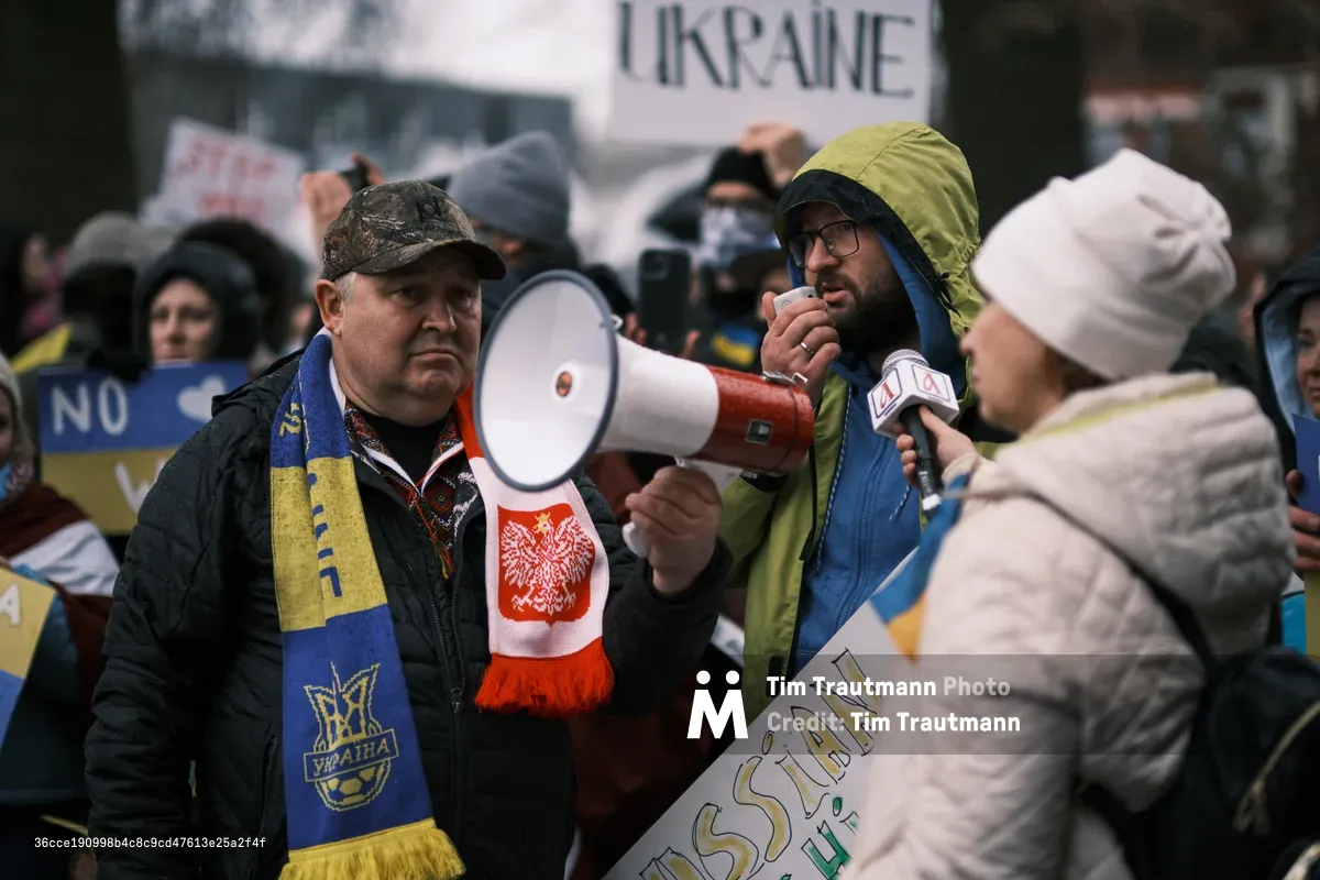 In the gray winter light outside Portland's Revolution Hall, protesters gather in solidarity with Ukraine as tensions mount. A man in camouflage cap and blue-yellow Ukrainian scarf speaks passionately into a red megaphone, his breath visible in the cold air. Around him, bundled demonstrators hold signs reading "UKRAINE" while a reporter captures testimonies of those calling for peace in the days before conflict would erupt.