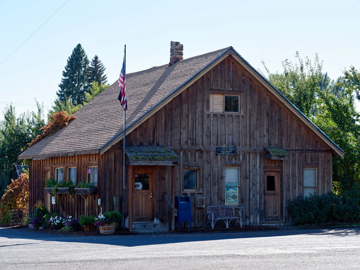 A weathered wooden post office building stands as a testament to small-town America in Glenwood, Washington. The rustic cedar-shingled structure features moss-covered eaves and a prominent American flag, creating an atmospheric portrait of rural postal service. Soft evening light bathes the scene, highlighting the building's authentic patina and the carefully tended flower boxes that add touches of color to the weathered facade. The composition captures both the utilitarian purpose and enduring charm of this community cornerstone nestled among towering evergreens.