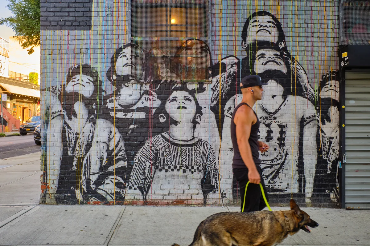 Golden hour light bathes a powerful mural on South 6th Street in Williamsburg, Brooklyn, featuring monochromatic portraits of cultural figures rendered in striking black and white against weathered brick. A man in athletic wear walks his German Shepherd past the towering artwork, creating a compelling juxtaposition between monumental street art and intimate neighborhood moments. The warm evening glow illuminates both the vibrant paint drips cascading down the wall and the peaceful domestic scene unfolding below.
