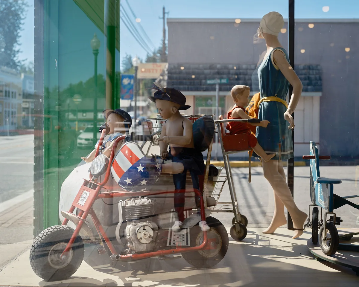 A quirky antique or thrift store window display in Vernonia, Oregon, photographed through the glass with the sunny small-town street reflected in it. The display features several vintage child mannequins posed on and around a red mini motorcycle with an American flag-patterned fairing. A female adult mannequin in a teal 1920s-style dress and white cap pushes a vintage metal shopping cart with a child mannequin seated in it. A small vintage tricycle sits to the right. The main street of Vernonia, including a green lamppost, utility poles, and storefronts, is visible in the reflection.