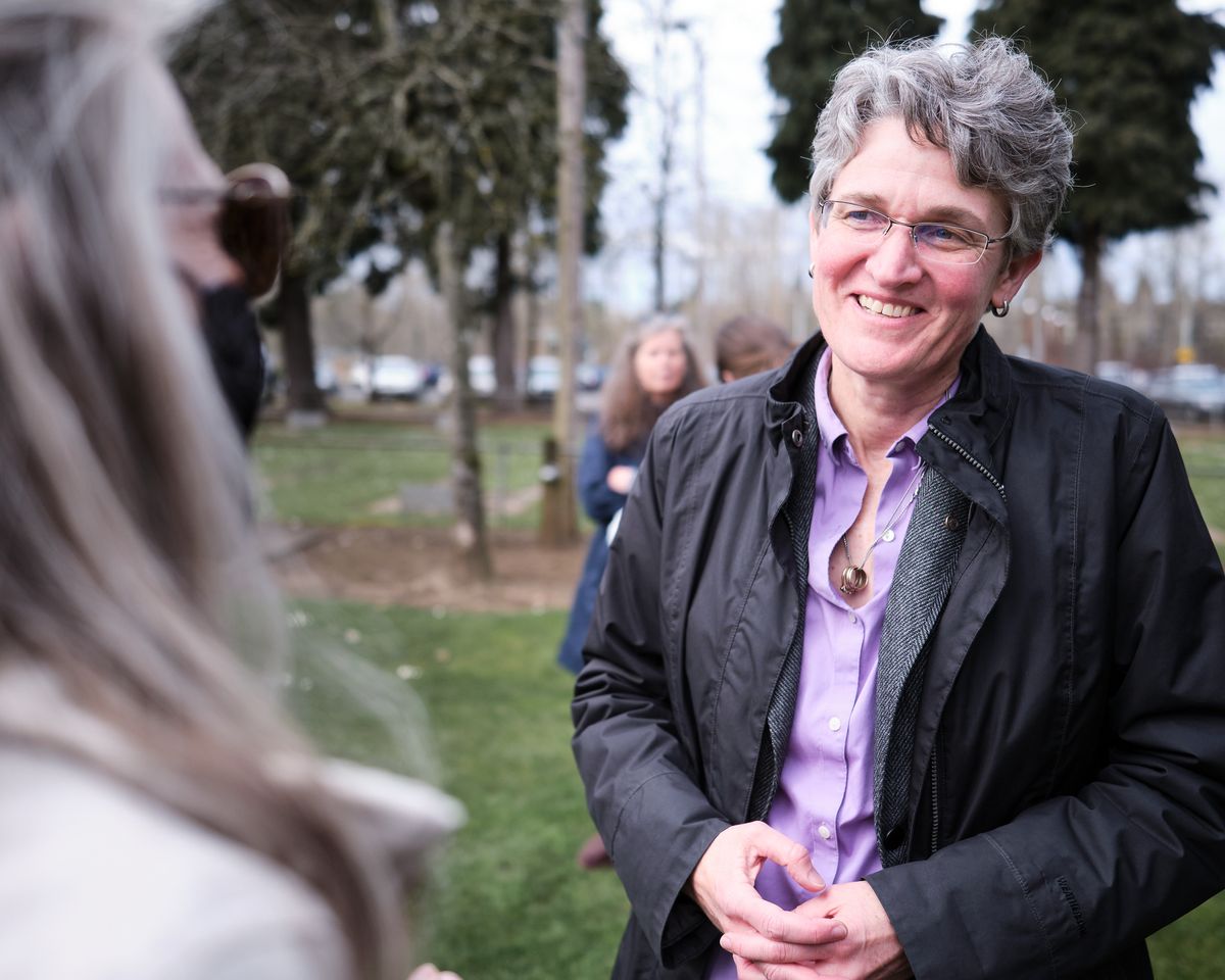 A middle-aged woman with gray curly hair and glasses smiles warmly while engaged in conversation in a park setting. She wears a black jacket over a purple shirt, standing in what appears to be a public space in Oregon City with trees and other people visible in the blurred background.