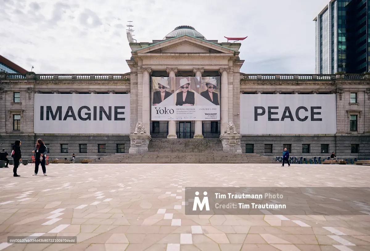 The neoclassical facade of the Vancouver Art Gallery stands transformed into a canvas for peace, with Yoko Ono's iconic "IMAGINE PEACE" message spanning its limestone columns. Under a gentle overcast sky, the building's grand pediment frames a promotional banner for Ono's exhibition, while scattered visitors cross the weathered stone plaza. The juxtaposition of classical architecture with contemporary art activism creates a powerful statement in the heart of downtown Vancouver's cultural district.
