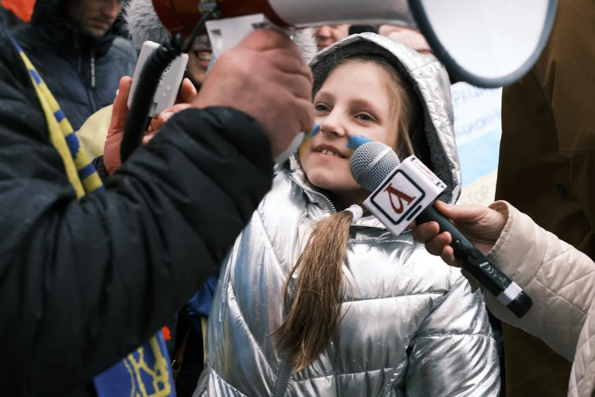 A young woman in a metallic silver puffer jacket becomes the focal point of an intense media gathering outside Portland's Revolution Hall, her face illuminated by the convergence of microphones and recording devices. Blue face paint streaks across her cheeks as journalists and activists press forward, creating a claustrophobic swirl of winter coats and extended arms. The harsh lighting and tight framing capture the raw energy of grassroots activism meeting mainstream media coverage. Behind her, the blurred presence of other protesters suggests the broader movement gathering momentum in the days before international conflict would escalate.