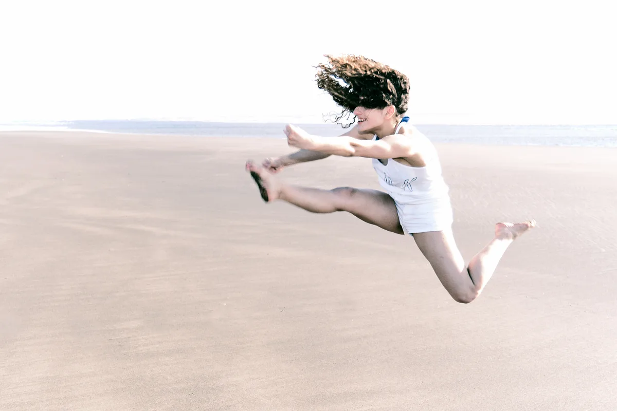 A young woman leaps joyfully into the air on a wide, flat sandy beach, her curly dark hair flying across her face. She wears a white tank top and shorts, arms outstretched and legs bent mid-jump, laughing as she soars above the sand. The bright, overcast sky and pale beach stretch to the horizon behind her.