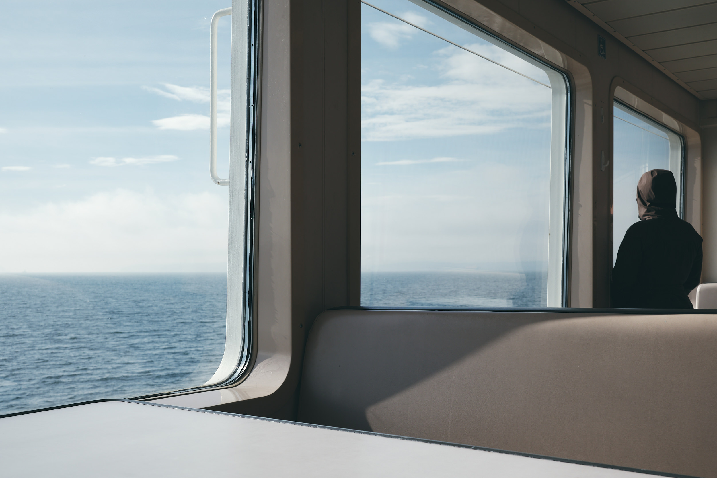 A person on a Washington State ferry is looking out of a window overlooking the ocean and its horizon. The ferry's seating accomodations are visible in the foreground.