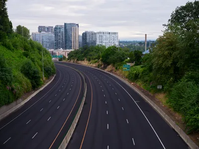 A wide, empty multi-lane highway curves through lush green hills toward the modern high-rise buildings of downtown Portland, Oregon under an overcast sky. The scene captures the contrast between urban development and natural landscape typical of the Pacific Northwest.