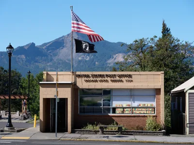 The mid-century brick and concrete United States Post Office stands sentinel in Cascade Locks, Oregon, its modernist lines softened by the dramatic backdrop of forested mountains rising into crystal blue sky. Two flags—the Stars and Stripes and a black POW/MIA banner—flutter from a single pole, while vintage street lamps frame the humble federal building. The Columbia River Gorge's towering peaks create a theatrical backdrop for this essential piece of small-town American infrastructure, where community connection meets governmental service.