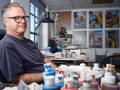 A middle-aged man with glasses and gray hair sits in his art studio surrounded by colorful paint tubes, brushes, and artwork displayed on the walls. The bright, organized workspace features large windows, a desk lamp, and various artistic materials scattered throughout the creative environment.