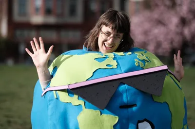 A jubilant woman wearing wire-rimmed glasses emerges from an oversized Earth costume, her arms raised in triumphant celebration against the soft-focus backdrop of Portland's Revolution Hall. The handcrafted globe features vivid azure oceans and lime-green continents, while a distinctive pink laptop computer sits incongruously across the equator. Golden afternoon light illuminates her genuine smile and the costume's felt textures, creating an atmosphere of playful environmental activism in Oregon's Central Eastside district.