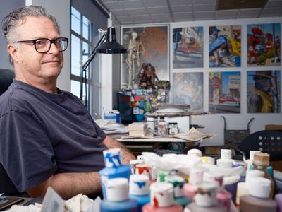 A middle-aged man with glasses and gray hair sits in his art studio surrounded by colorful paint tubes, brushes, and artwork displayed on the walls. The bright, organized workspace features large windows, a desk lamp, and various artistic materials scattered throughout the creative environment.
