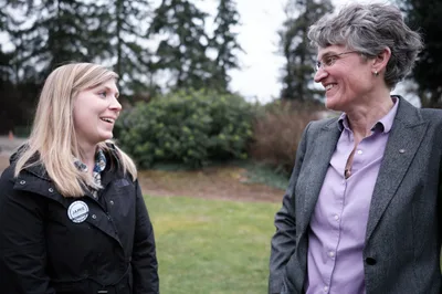Two women stand facing each other in an outdoor park setting in Oregon City, Oregon, engaged in friendly conversation and smiling. The younger blonde woman on the left wears a dark jacket with a campaign button, while the older woman on the right wears glasses, a gray blazer, and purple shirt.