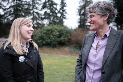 Two women stand facing each other in an outdoor park setting in Oregon City, Oregon, engaged in friendly conversation and smiling. The younger blonde woman on the left wears a dark jacket with a campaign button, while the older woman on the right wears glasses, a gray blazer, and purple shirt.