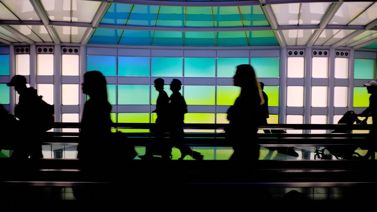 Travelers are silhouetted against the vivid backlit art installation "The Sky's the Limit" in the underground pedestrian tunnel connecting terminals at Chicago O'Hare International Airport. The curved ceiling and wall panels glow in shifting bands of turquoise, green, and yellow, creating a striking contrast against the dark figures of passing passengers. Their reflections shimmer on the polished floor below.