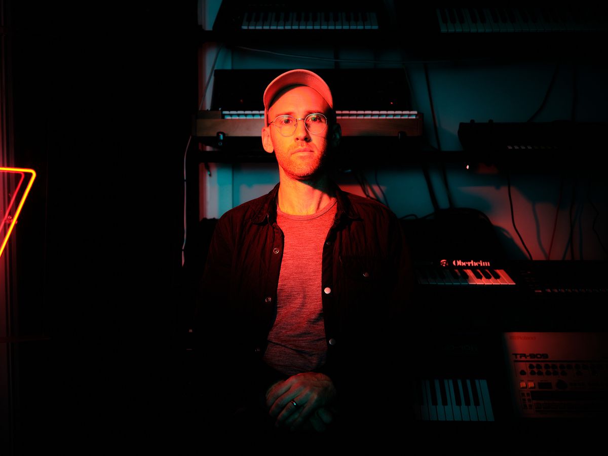 A portrait of DJ and event producer Peter Marks seated in a darkened music studio, lit dramatically by warm red light. He wears round glasses, a baseball cap, a grey t-shirt, and an open dark shirt. An Oberheim synthesizer and a Roland TR-909 drum machine are visible in the foreground, with additional keyboards and equipment stacked behind him. A red neon triangle glows on the left side of the frame.