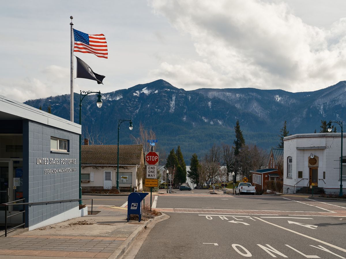 The United States Post Office in Stevenson, Washington, a single-story blue brick building with signage reading "Stevenson Washington 98648." An American flag and a black POW/MIA flag fly from a tall flagpole. A blue USPS mailbox sits on the sidewalk near a stop sign and "End One Way" sign at an intersection. In the background, a quiet small-town main street leads toward the Columbia River Gorge, with densely forested mountains dusted with snow rising dramatically under a partly cloudy sky.