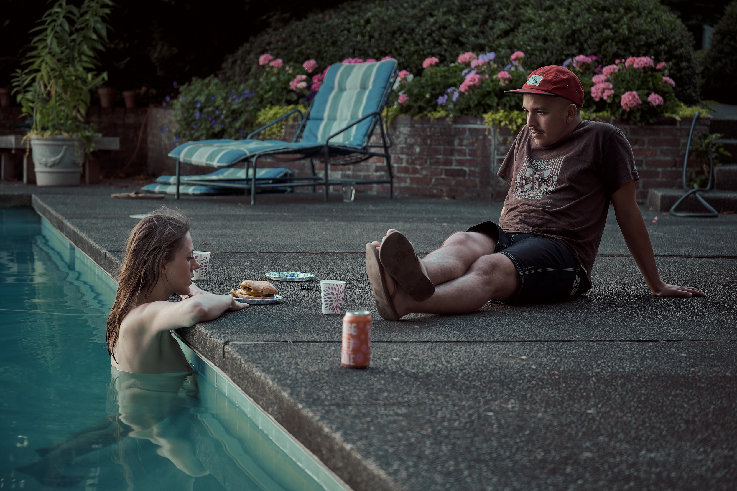 At dusk, a woman with wet hair rests her arms on the edge of a backyard swimming pool, chatting with a man who sits on the pool deck beside her. Between them sit plates of food, patterned paper cups, and a canned drink. The man wears a red baseball cap, a brown t-shirt, and dark shorts. A turquoise lounge chair, brick retaining wall, and lush pink flowering shrubs are visible in the softly lit background.