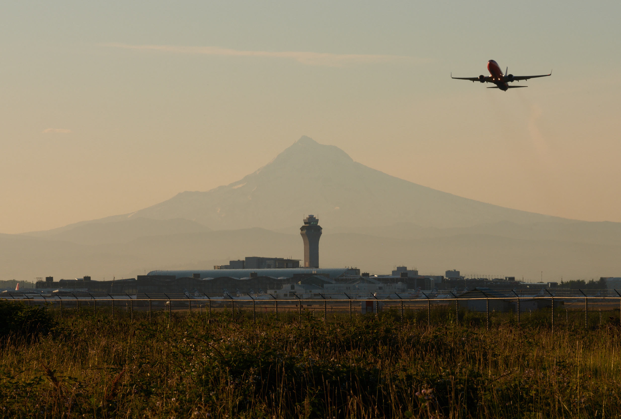 A commercial aircraft flies through a hazy sky above an airport control tower and terminal buildings, with a large mountain silhouetted in the background during golden hour lighting. The foreground shows wild grass and vegetation with a security fence marking the airport perimeter.