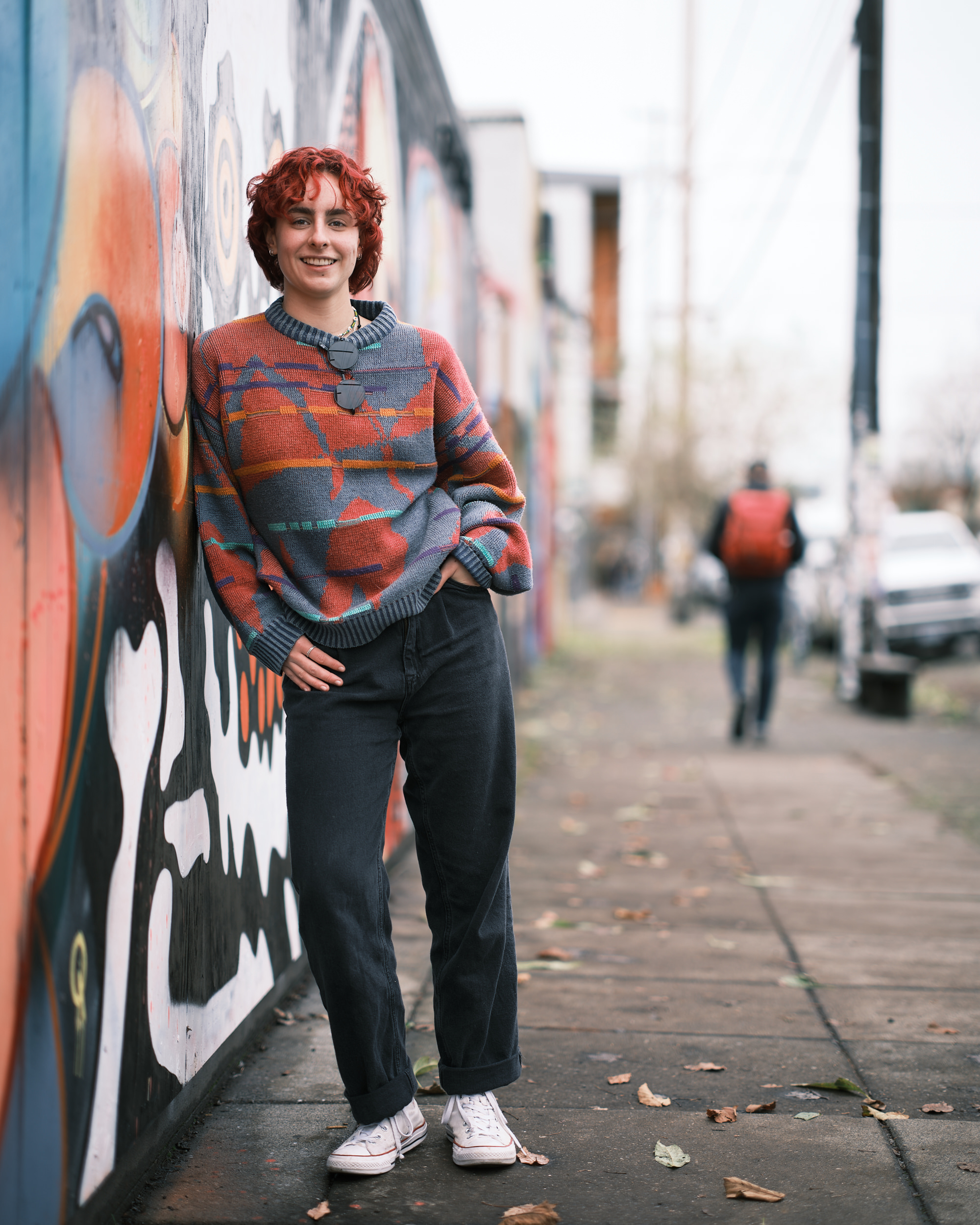 A full-length senior portrait of Annika Molina leaning against a colorful street mural on a Portland sidewalk. She has short curly red hair and wears an oversized patterned sweater in orange, grey, and teal, dark wide-leg jeans cuffed at the ankle, white Converse sneakers, and a pendant necklace. She smiles warmly at the camera with one hand on her hip. A pedestrian with a red backpack walks in the blurred background along the tree-lined street.