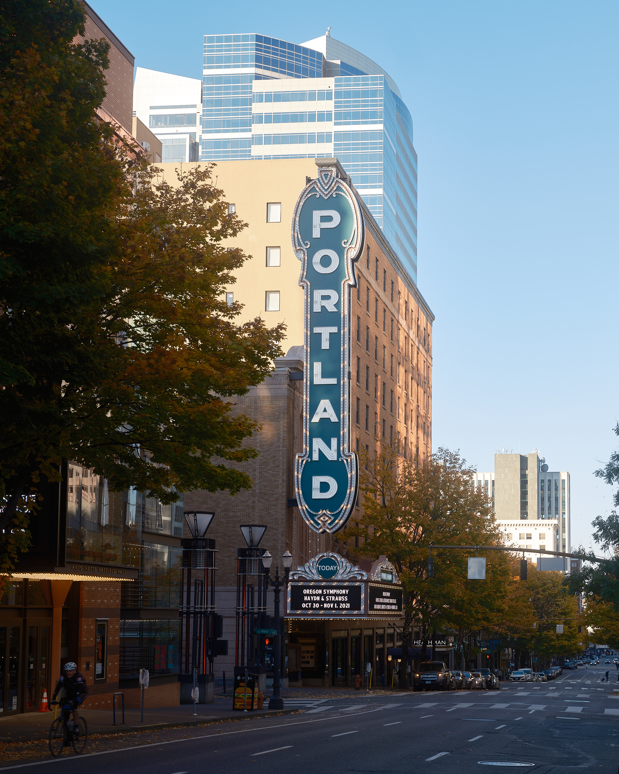 The exterior of the Arlene Schnitzer Concert Hall on Broadway in downtown Portland, Oregon, photographed on a clear autumn day. The venue's iconic large teal and white vertical "Portland" marquee sign dominates the facade of the historic brick building. The theater's marquee below reads "Oregon Symphony — Haydn & Strauss — Oct 30 - Nov 1, 2021." Autumn trees in golden and orange foliage frame the street, and a cyclist rides past in the foreground. The Heathman Hotel is visible further down the block, with modern glass office towers rising behind the historic building against a clear blue sky.