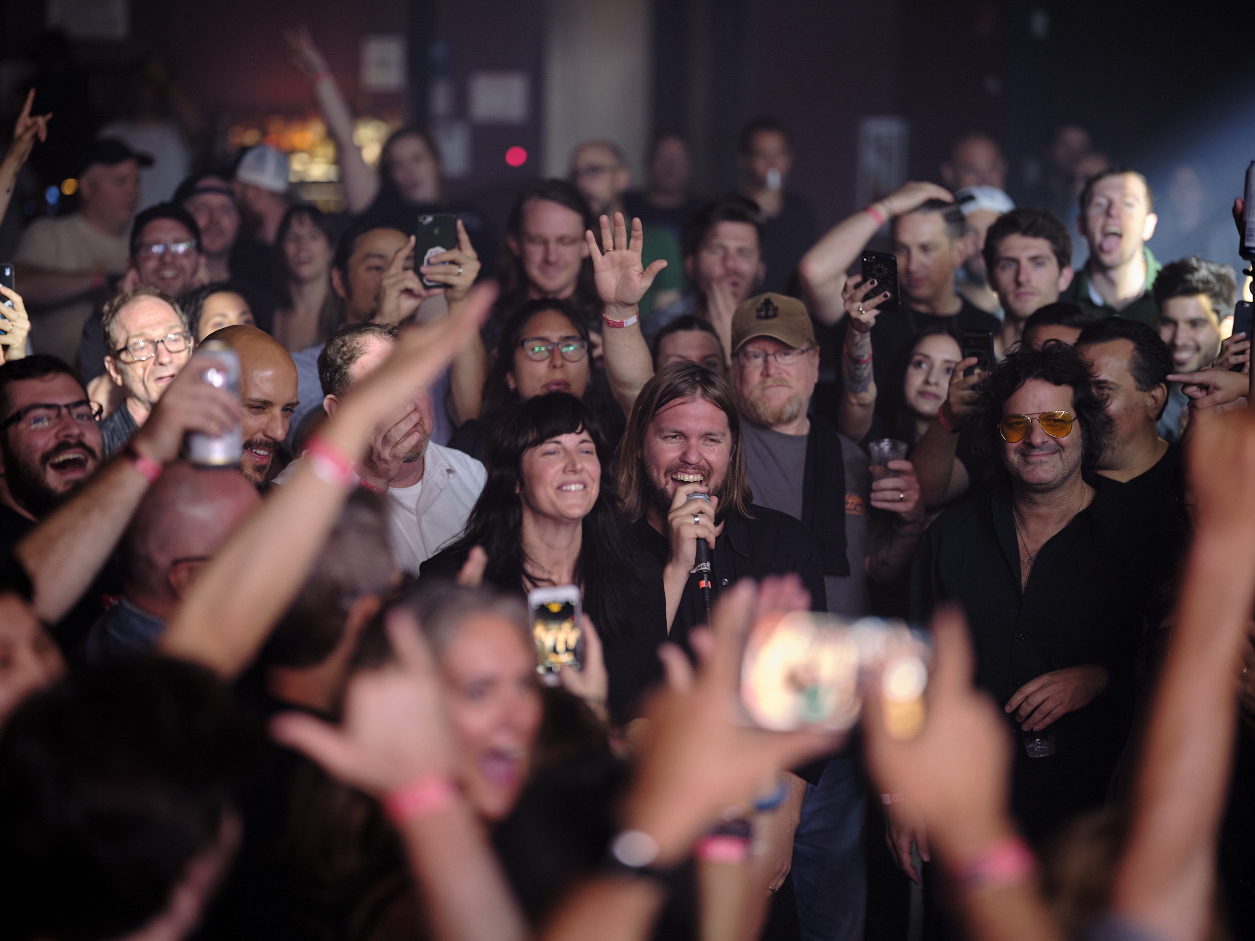 Band of Skulls members Matt Hayward and Emma Richardson perform in the middle of a packed, energetic crowd at Webster Hall in New York City in September 2019. Matt, with long hair and wearing a black shirt, holds a microphone and smiles broadly, while Emma stands beside him. Audience members surrounding them cheer, raise their hands, and hold up smartphones to capture the moment. The dimly lit venue is filled with excitement and motion blur in the foreground.