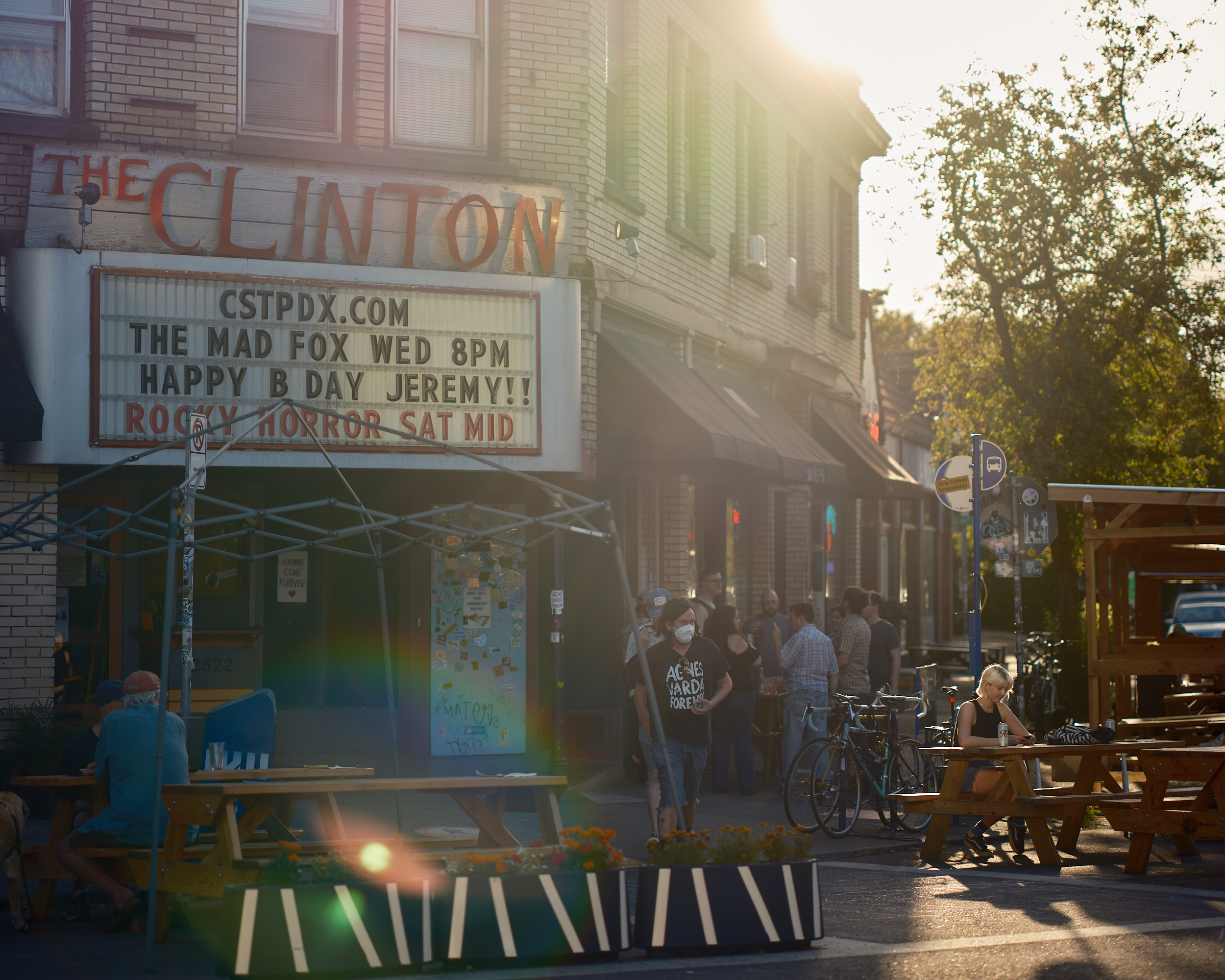 Alt text:  The exterior of The Clinton Street Theater in Portland, Oregon, bathed in warm golden lens-flare sunlight. The theater's marquee reads "The Mad Fox Wed 8PM — Happy B Day Jeremy!! — Rocky Horror Sat Mid." A lively crowd of people gathers on the sidewalk outside, with outdoor picnic tables and planters with marigolds occupying a parklet in the foreground. A woman in a face mask wearing a black "Agnès Varda Forever" t-shirt walks past. Bicycles are parked along the curb, and a woman sits alone at a picnic table to the right.