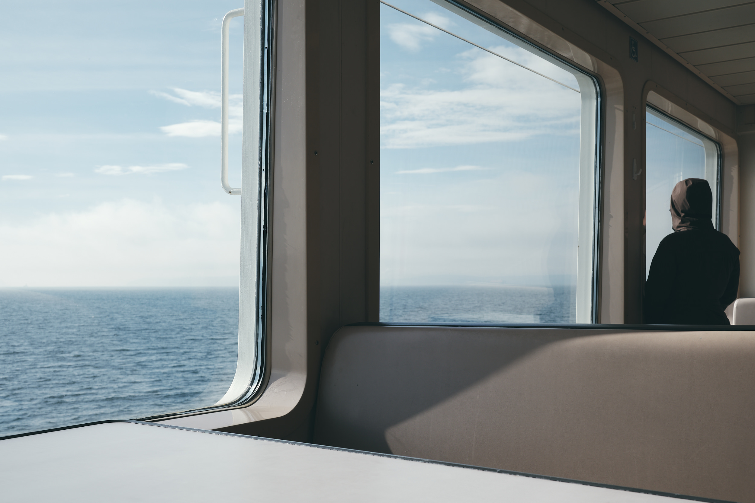 The interior of a Washington State Ferry passenger cabin, with large windows looking out over the calm blue waters of Puget Sound under a partly cloudy sky. A lone passenger wearing a dark jacket and head covering sits with their back to the camera, gazing out at the open water. Empty beige bench seating runs along the window row in the foreground.