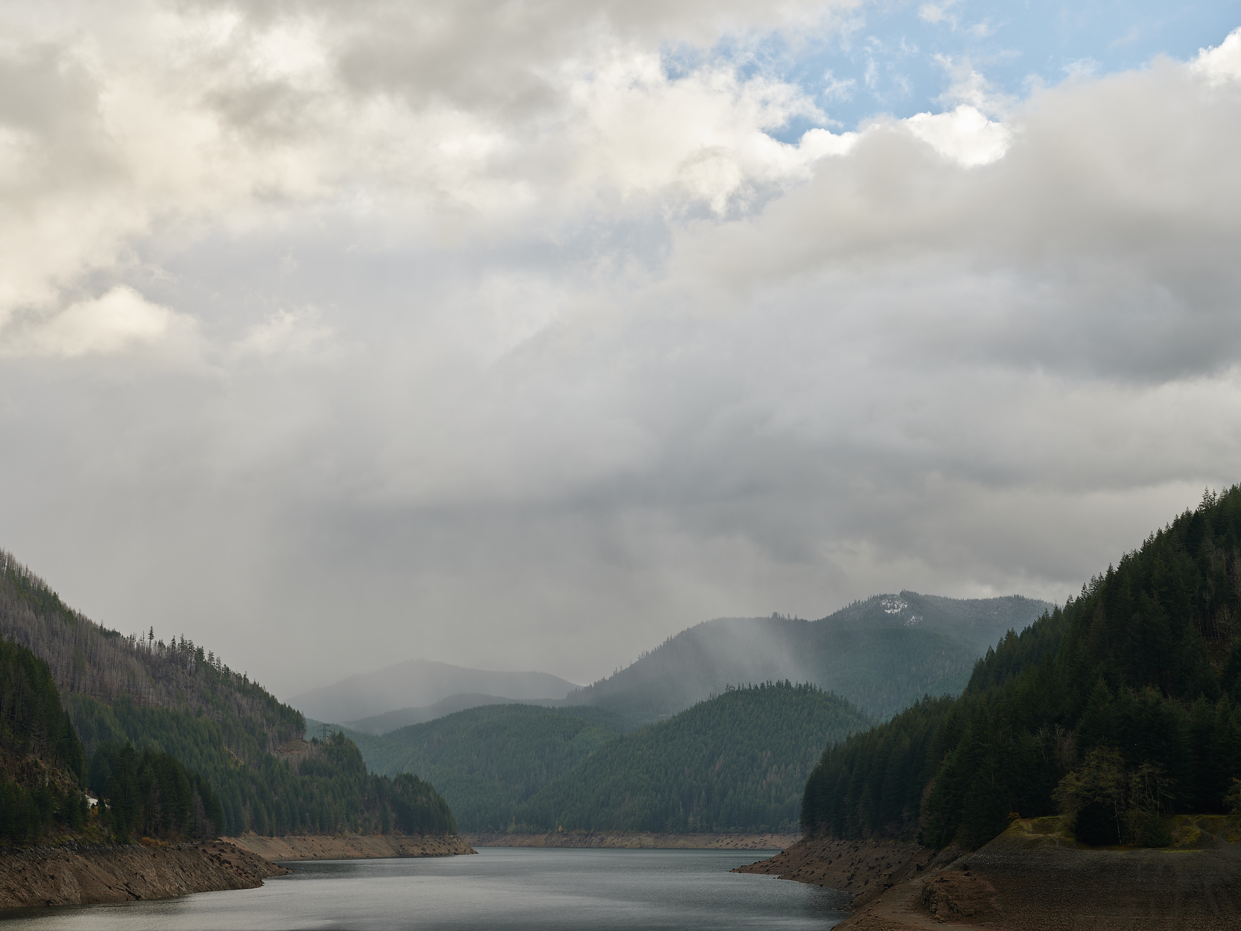 This is a moody, atmospheric landscape shot of Detroit Lake in Oregon. The reservoir stretches into the middle distance, flanked on both sides by steep, densely forested hillsides of Douglas fir and pine. The water level appears notably low, with exposed rocky and muddy shoreline visible along the banks - likely reflecting seasonal drawdown or drought conditions.  The surrounding hills show a mix of healthy green conifers and what appear to be fire-damaged or dead trees on the left slope, a reminder of the wildfires that have affected this region in recent years.  The sky dominates the upper two-thirds of the frame and is dramatic — heavy, layered storm clouds roll in from the left, with soft light breaking through on the right side, hinting at blue sky barely visible above. Mist or light rain obscures the middle mountains in the background, giving the scene a brooding, Pacific Northwest character.  The overall palette is cool and muted - slate grays, deep forest greens, and earthy browns - which gives the photo a quiet, contemplative feel. It looks like it was taken from the dam or a viewpoint above the lake, offering a clear line of sight straight up the reservoir corridor.