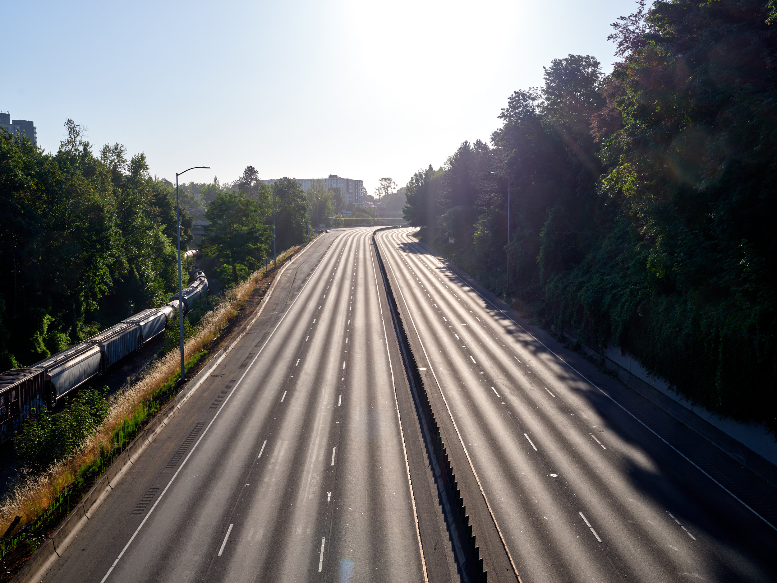 An aerial view of the empty Banfield Freeway (Interstate 84) in Portland's Lloyd District, showing multiple lanes of asphalt stretching into the distance with lush green trees lining both sides and urban buildings visible in the background under bright sunlight.