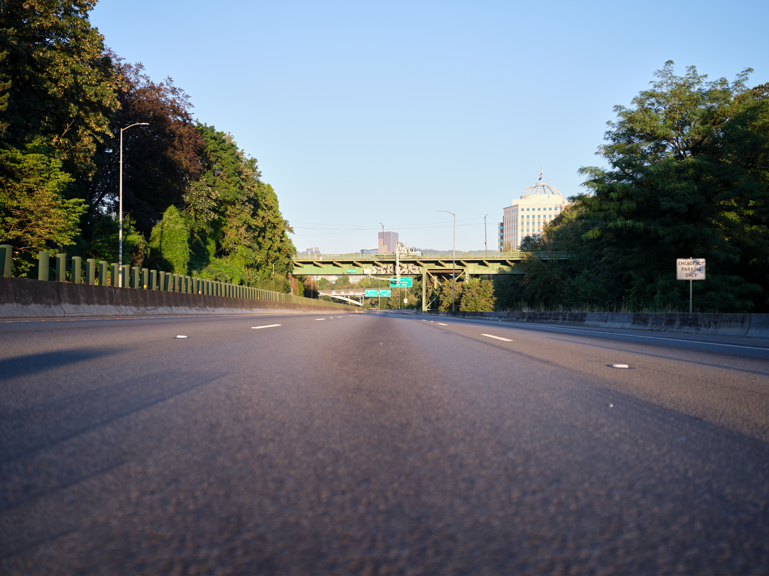 A low-angle view of the empty Banfield Freeway (I-84) in Portland's Lloyd District, showing the asphalt road surface with lane markings leading toward downtown buildings visible in the distance. Lush green trees line both sides of the highway, with a concrete overpass bridge crossing above and clear blue skies overhead.