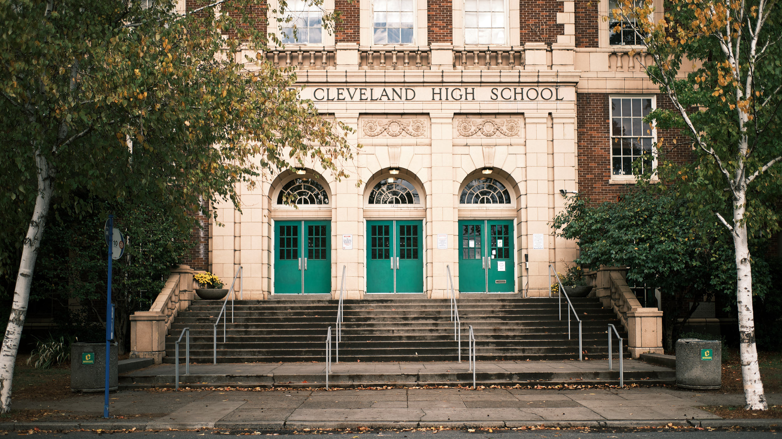 The stately historic facade of Grover Cleveland High School in Portland, Oregon, photographed on an autumn day. The building features a classical red brick and cream stone exterior with three arched entryways fitted with teal green double doors and fanlight windows above. "Cleveland High School" is carved in stone above the central entrance. Wide stone steps with metal handrails lead up to the three entrances. White-barked birch trees with yellowing autumn leaves frame the building on both sides, and fallen leaves scatter the sidewalk in the foreground.
