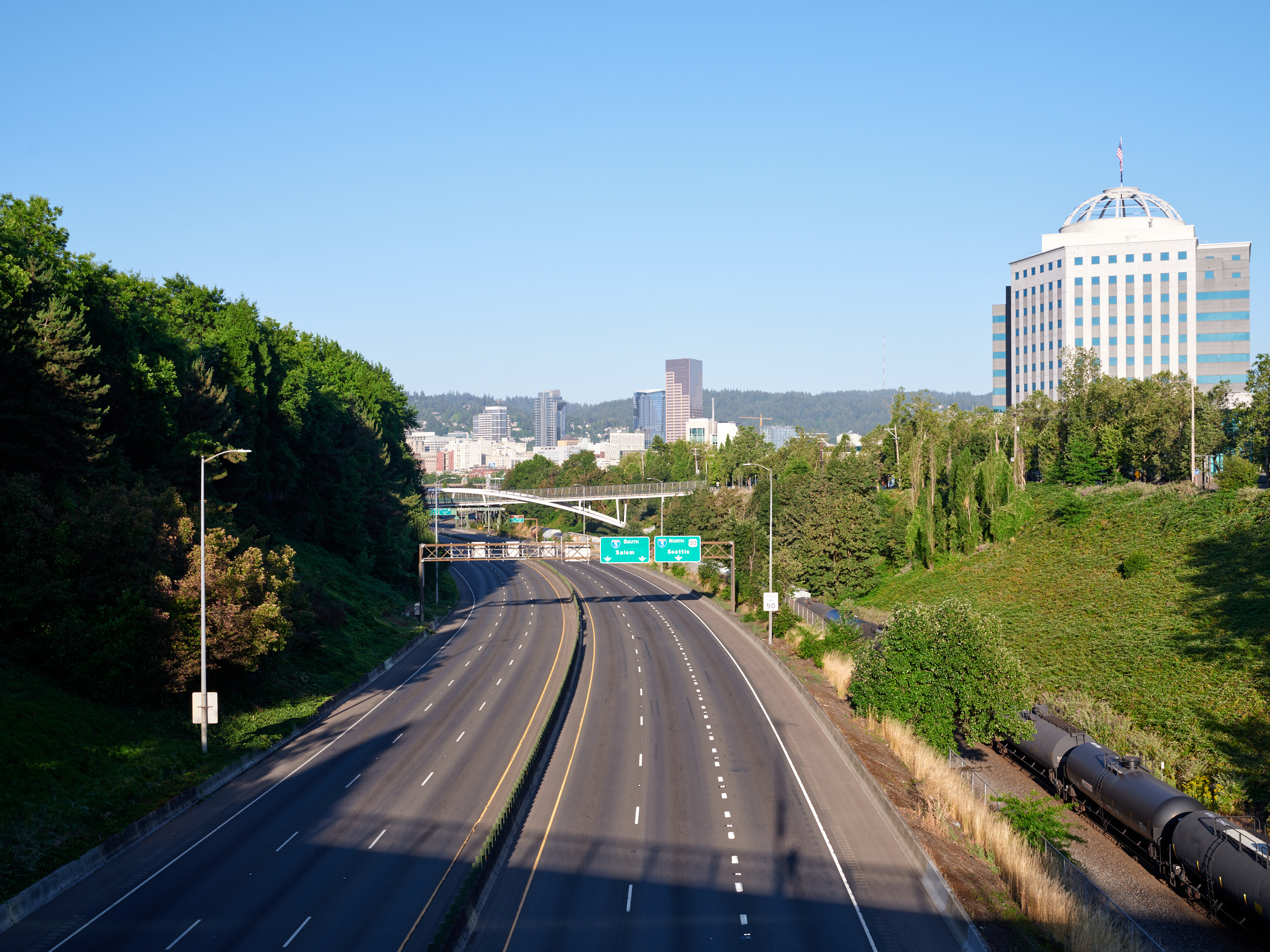 A multi-lane highway curves toward the downtown Portland, Oregon skyline, featuring modern office buildings and skyscrapers surrounded by lush green hills. The road is flanked by dense forests and includes green highway signs directing traffic toward the city center.