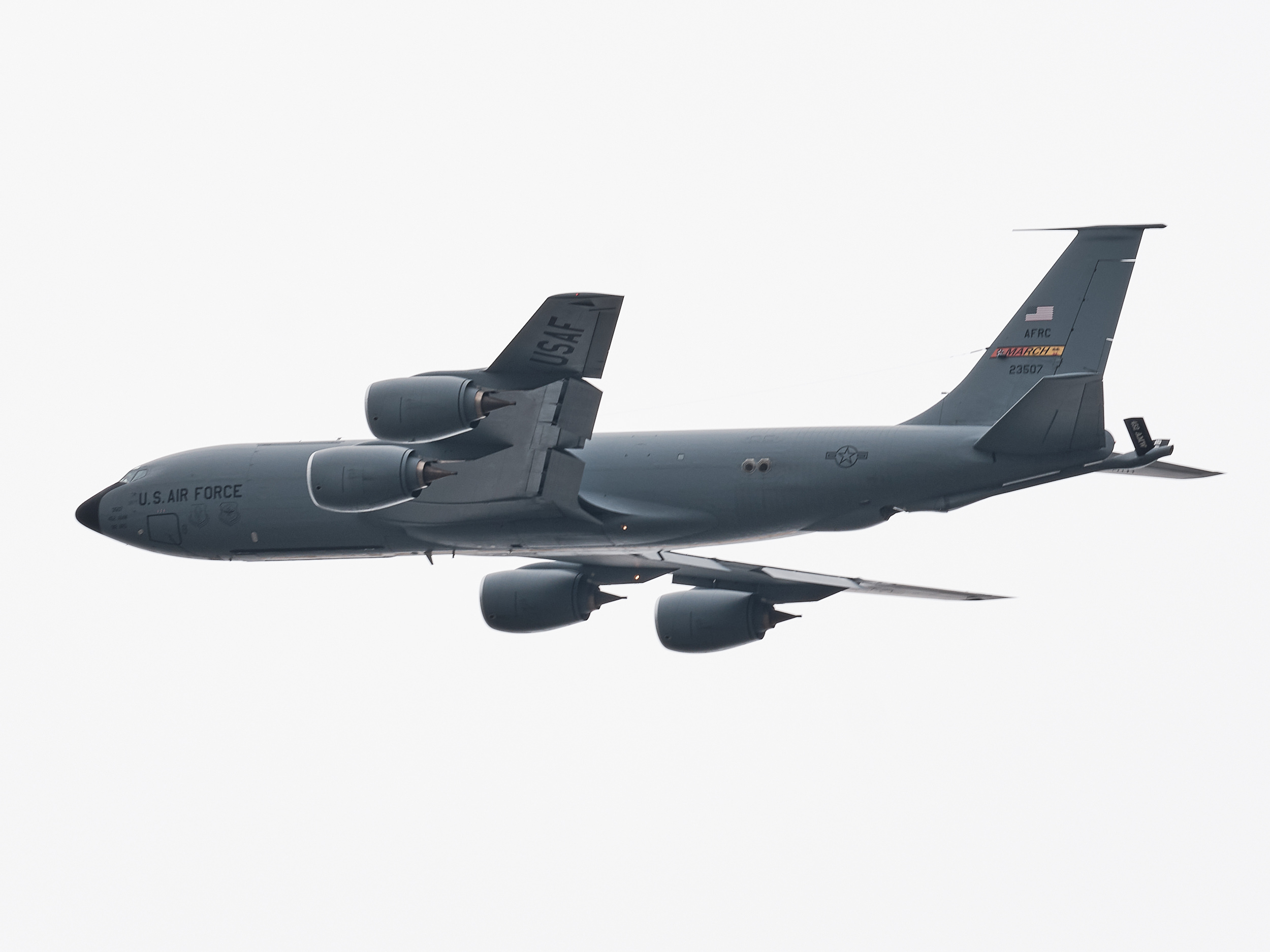 A U.S. Air Force Boeing KC-135 Stratotanker aerial refueling aircraft photographed in flight from below against a flat, overcast white sky. The aircraft is painted in overall grey and bears "U.S. AIR FORCE" lettering on the fuselage. "USAF" is stenciled on the vertical stabilizer, with "AFRC" and "MARCH" unit markings and the tail number 23507 visible near the tail. Four podded jet engines are mounted under the swept wings. The aircraft's boom refueling equipment is visible extending from the rear of the fuselage. A U.S. national insignia roundel appears on the fuselage. The tight telephoto framing isolates the aircraft cleanly against the grey-white sky.