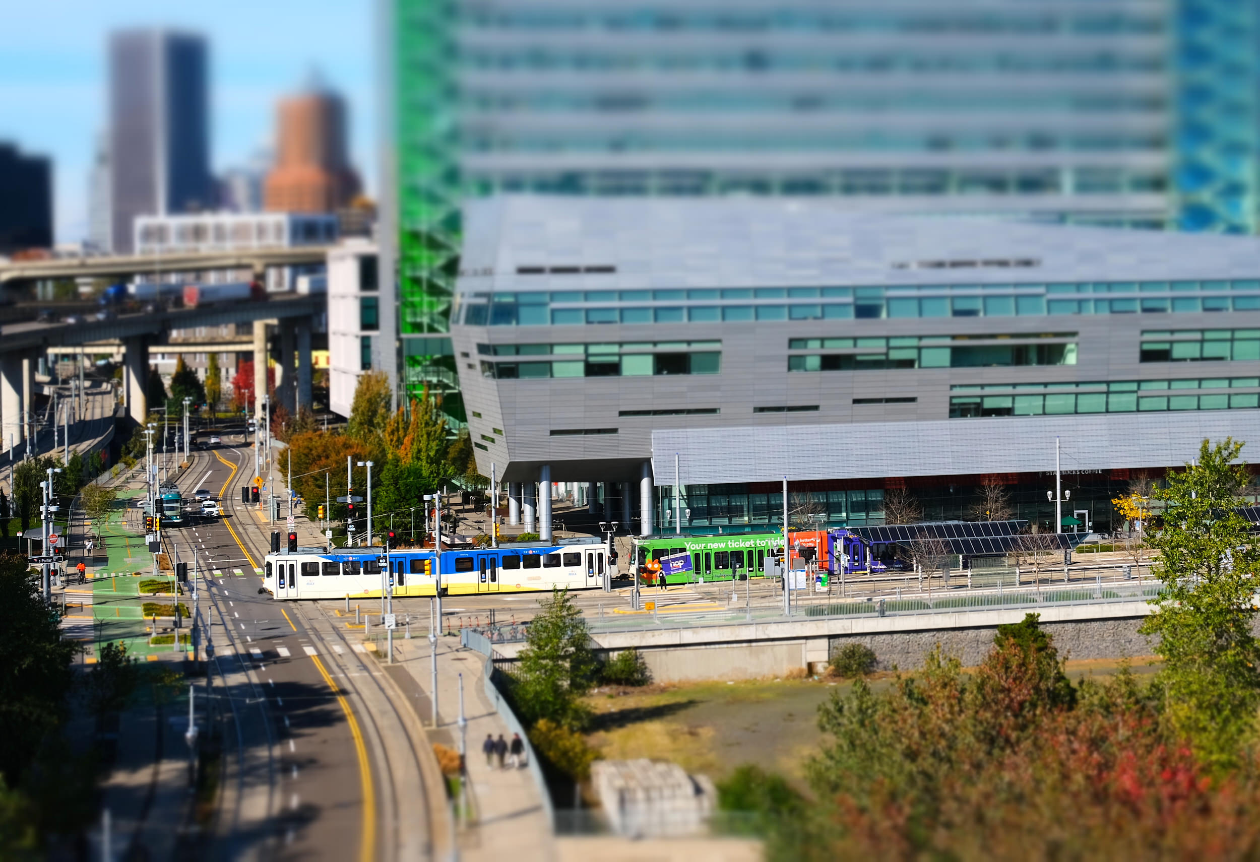 A tilt-shift photograph of Portland's South Waterfront district, giving the scene a miniature toy-like appearance. A TriMet MAX light rail train in blue and yellow livery runs along the street-level tracks in front of the OHSU Collaborative Life Sciences Building, a large modern structure clad in grey panels and green glass. A green-wrapped advertising train and a purple bus are visible at the adjacent transit stop. An elevated freeway interchange curves through the background, with the blurred Portland downtown skyline and autumn foliage visible in the distance under a clear blue sky.