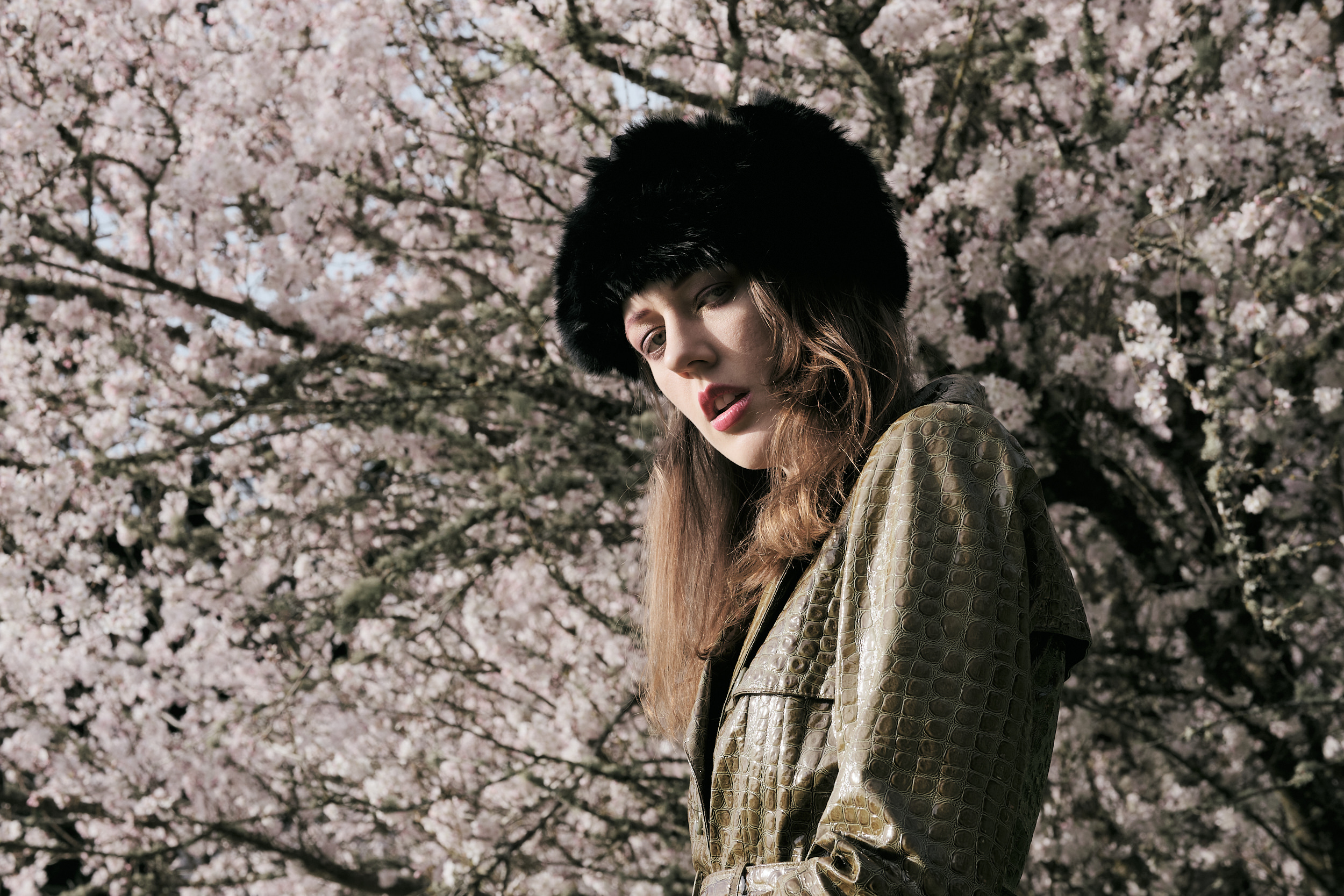 A portrait of Rhianna Atwood standing in front of a fully bloomed cherry blossom tree, its soft pink flowers filling the background. She wears an olive green crocodile-textured leather coat and a black fluffy fur hat, with long brown hair resting over her shoulders. She gazes downward with a serene expression in warm natural sunlight.