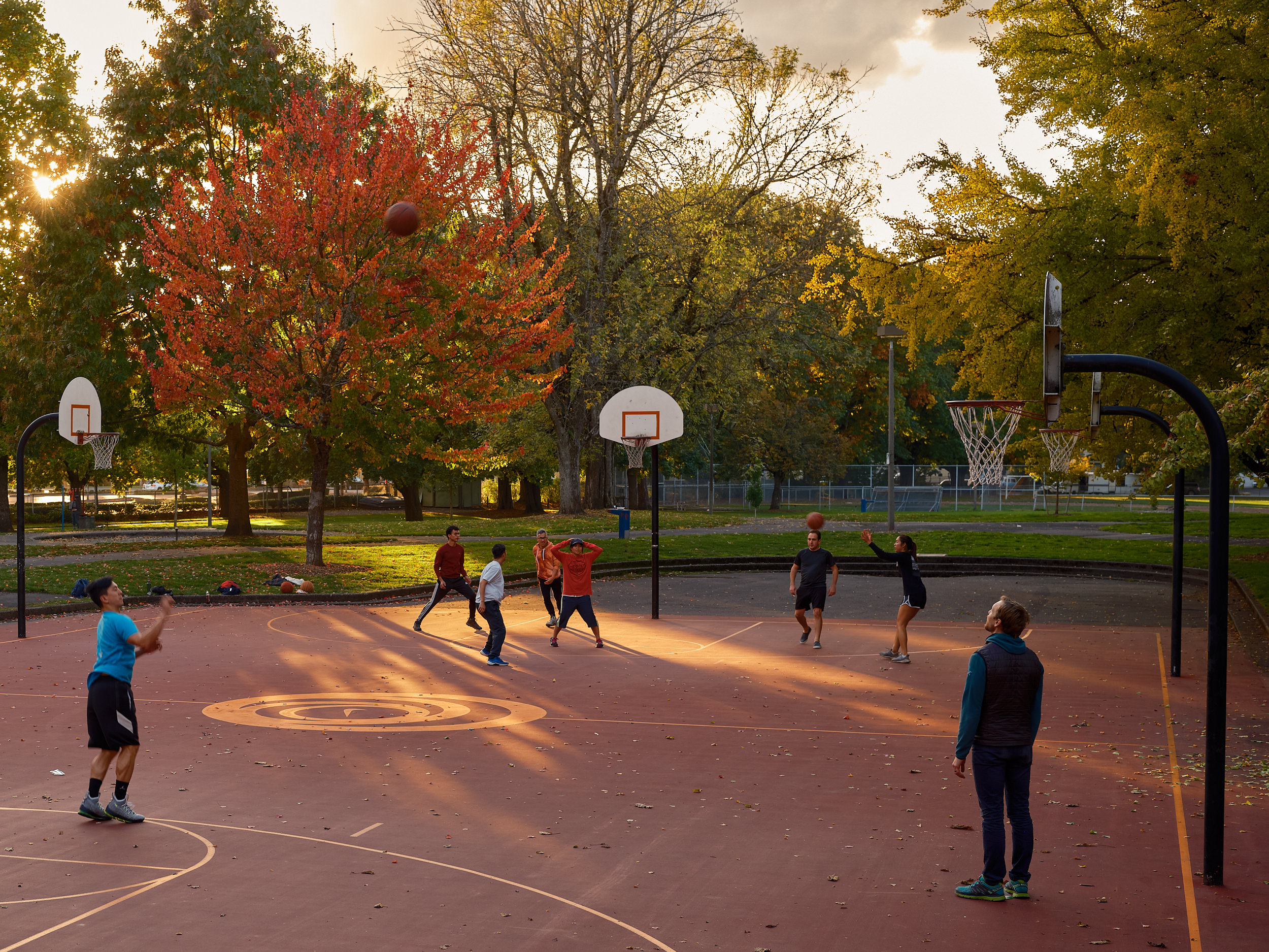 A group of people play pickup basketball on an outdoor court at Powell Park in Portland, Oregon, bathed in warm golden evening light. A basketball is suspended mid-air above the court. Three hoops are visible across the wide red-surfaced court, with fallen autumn leaves scattered across it. Vibrant fall foliage in shades of red, orange, and yellow fills the background, with the setting sun breaking through the trees on the left.