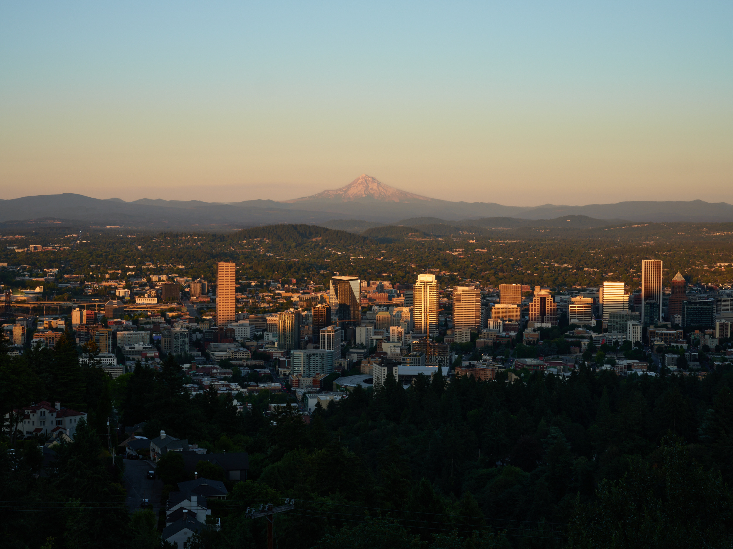 An elevated view of Portland, Oregon's downtown skyline at golden hour, with the snow-capped peak of Mount Hood visible in the distance beyond rolling hills. The city's high-rise buildings are illuminated by warm sunset light, while residential areas and dense forest cover the foreground and surrounding landscape.