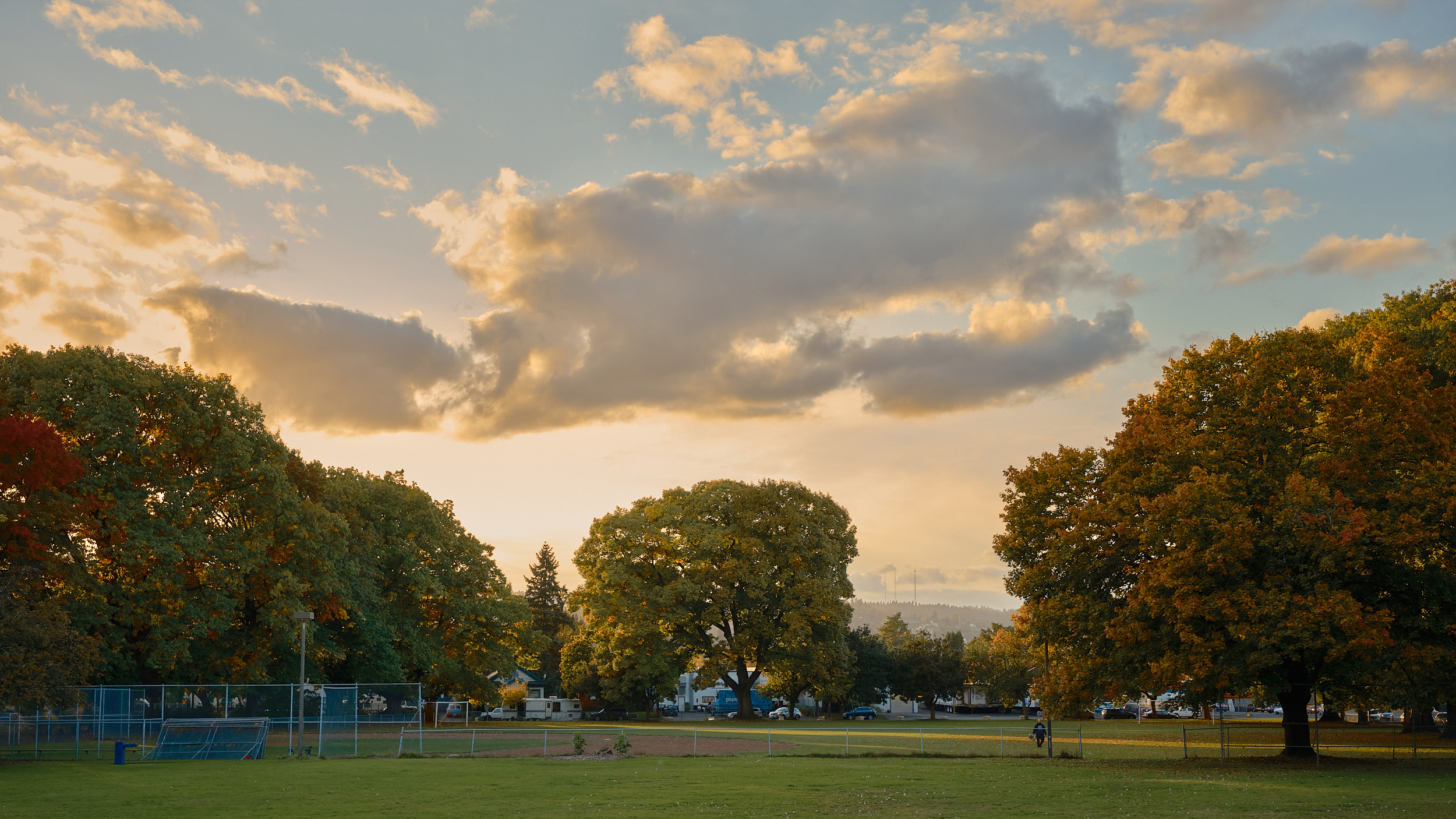 A sweeping autumn landscape at Powell Park in Portland, Oregon, bathed in warm golden evening light. Large mature trees in shades of green, amber, and rust frame an open grassy field with a baseball diamond and chain-link backstop visible on the left. A lone figure walks along the park's edge in the middle distance. Residential streets and parked vehicles are visible through the treeline, with the West Hills of Portland rising softly in the background. A dramatic sky of blue and gold-tinged clouds fills the upper half of the frame.