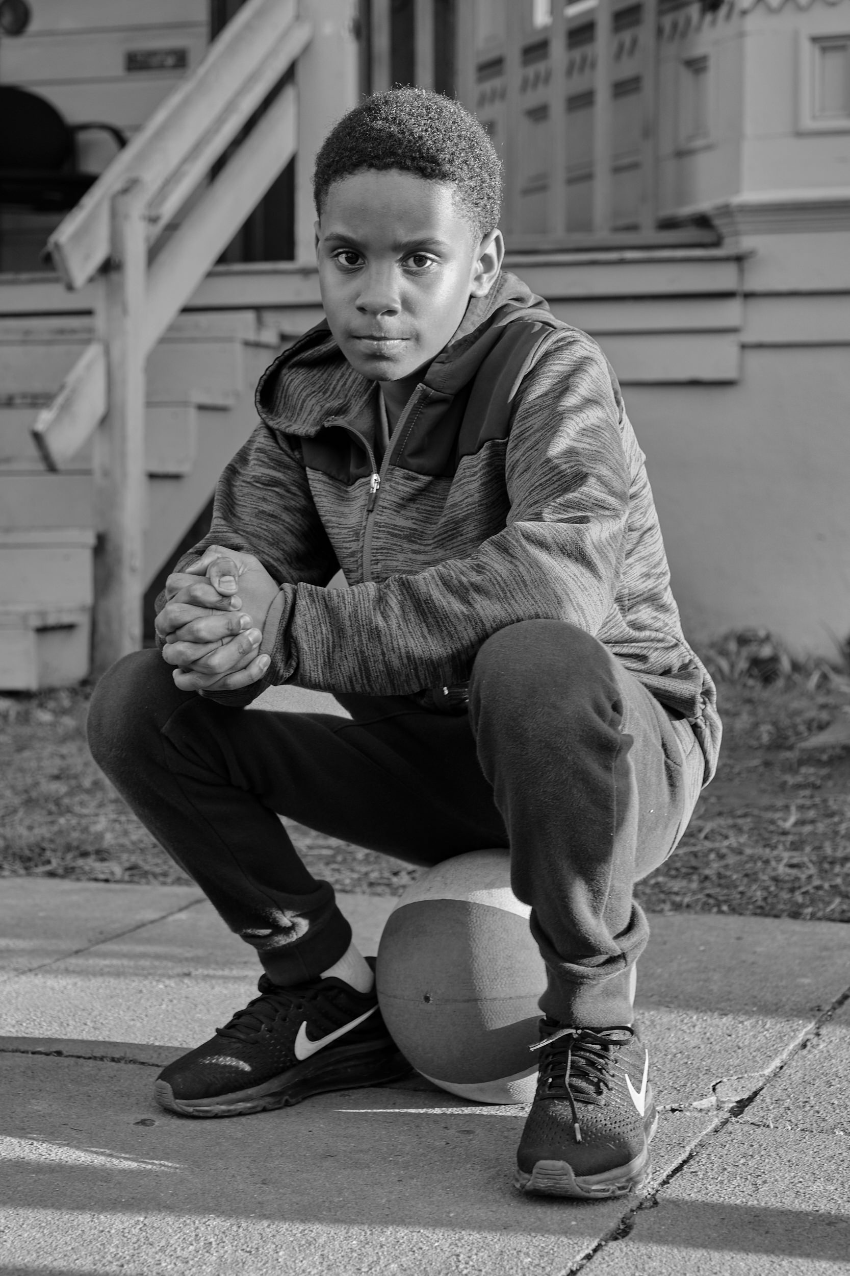 A black-and-white portrait of Kaye, a young boy, crouching on a sidewalk on Mississippi Avenue in Portland, Oregon. He wears a zip-up hoodie, sweatpants, and Nike sneakers, with his hands clasped together and resting on his knees. A basketball sits beneath his feet. He gazes directly into the camera with a steady, composed expression. A wooden porch and the facade of a residential house are visible in the background.
