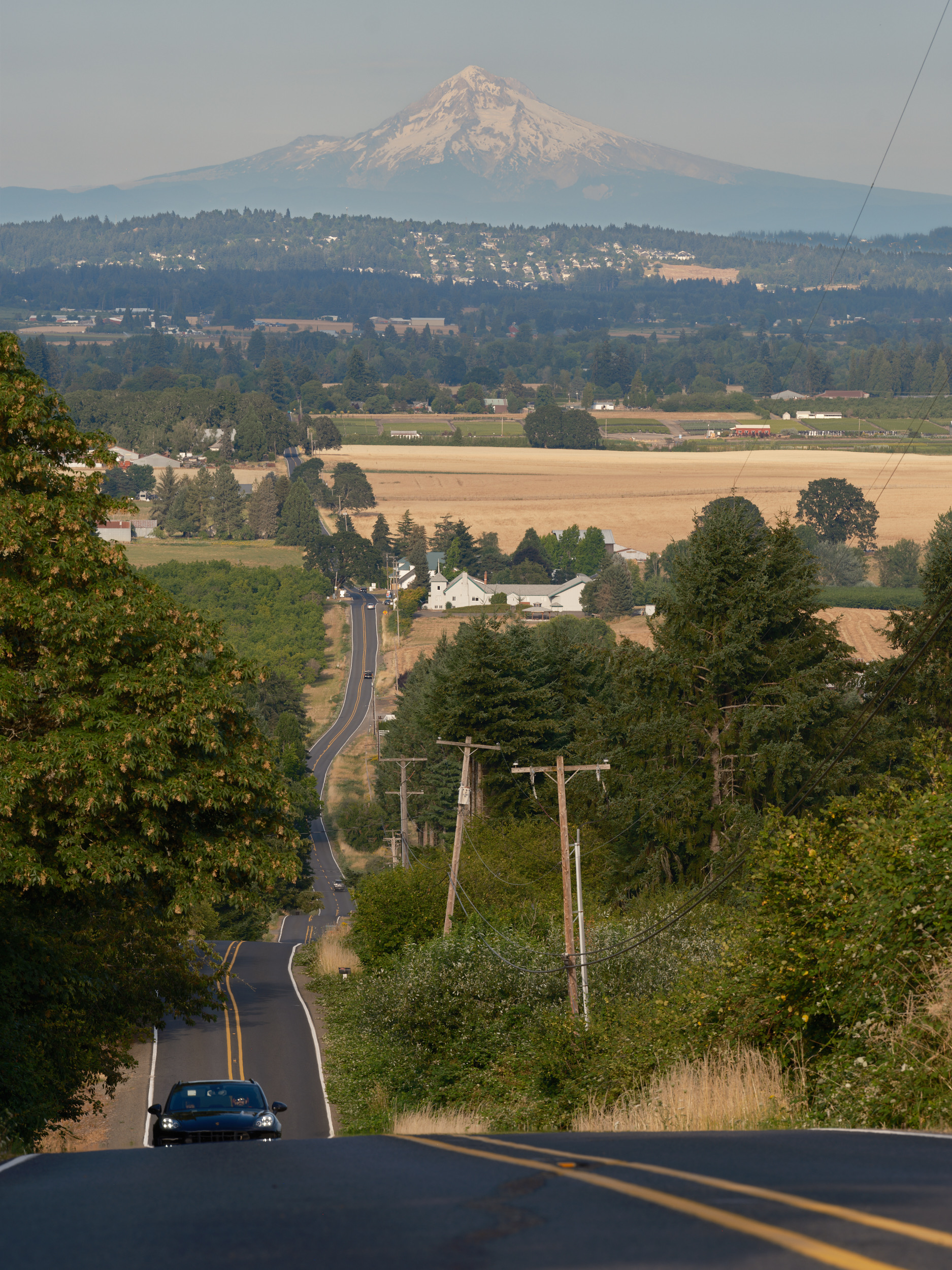A scenic rural road in Laurel, Oregon descends through rolling farmland with Mount Hood prominently visible in the background. Power lines run alongside the winding asphalt road, while a single car travels toward the camera through the pastoral landscape of golden fields and green trees.