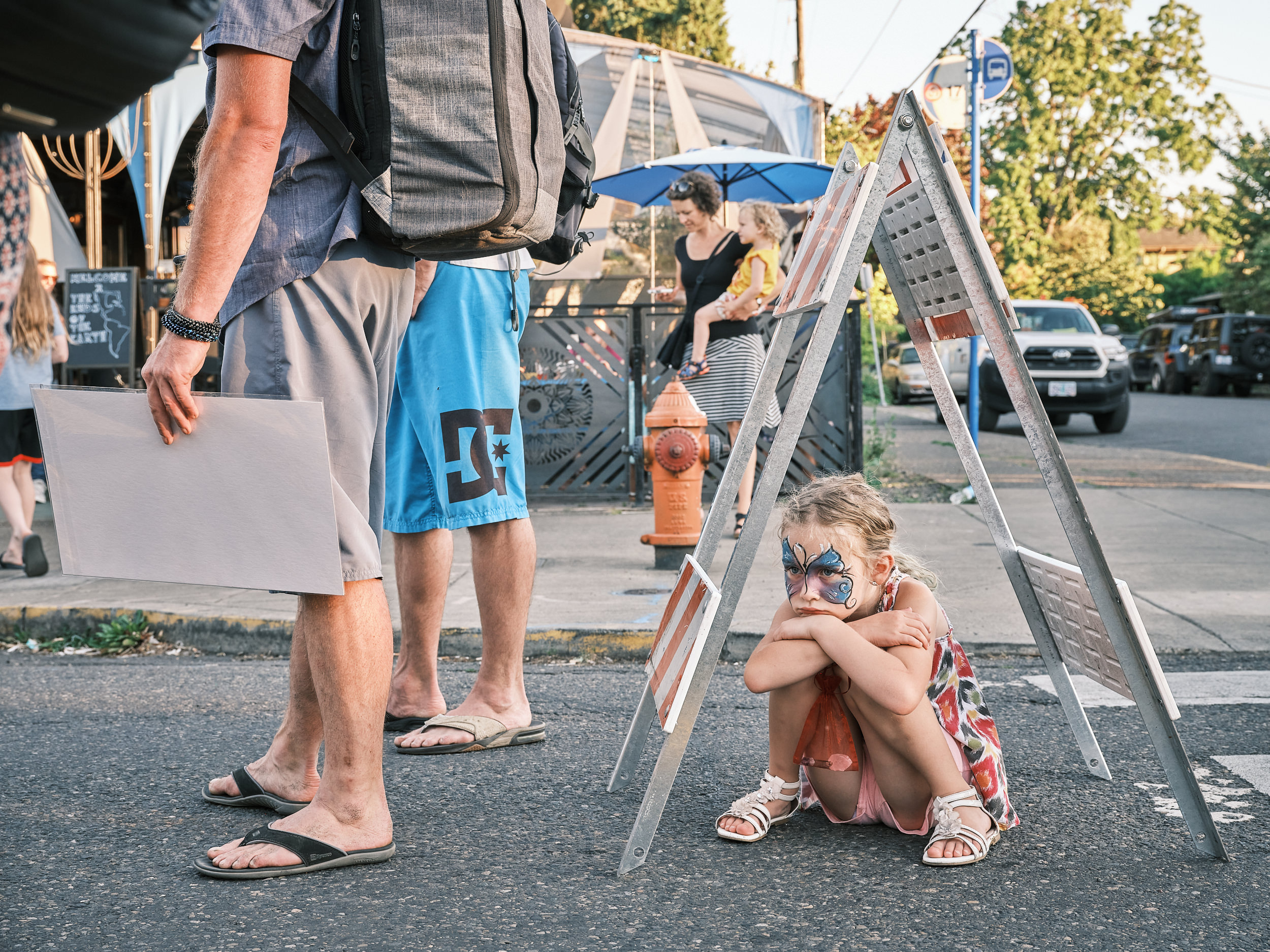 A young girl with a blue butterfly face painting sits curled up on the street beneath a metal A-frame ladder sign, hugging her knees with a sullen expression at the Last Thursday street fair on Alberta Street in Portland, Oregon. She wears a floral dress and white sandals. The legs of two adults standing nearby are visible in the foreground, and a woman holding a toddler is visible in the background near a blue patio umbrella and a red fire hydrant.