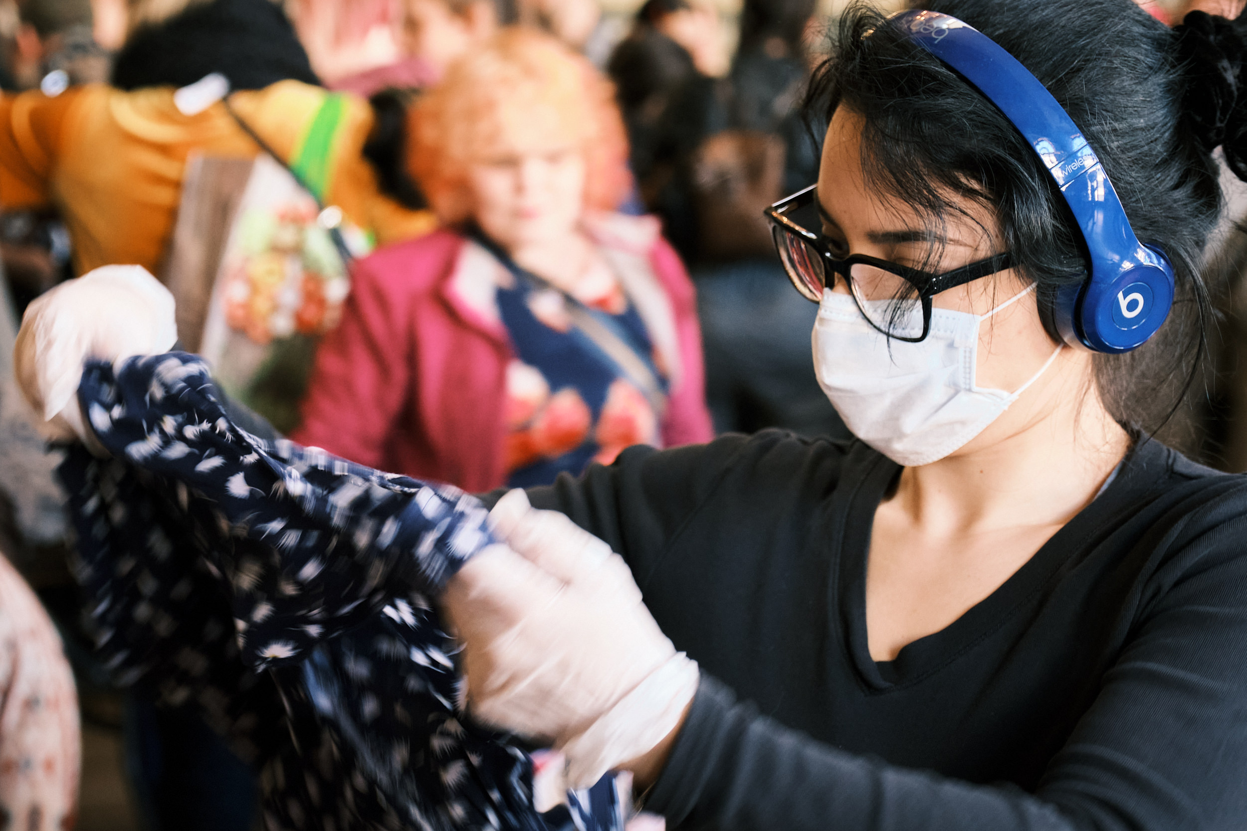A shopper wearing a white face mask, black-rimmed glasses, and blue Beats wireless headphones examines a patterned garment at a crowded swap meet inside the Crystal Ballroom in Portland, Oregon. She wears a black long-sleeve top and is focused on inspecting the clothing item. A busy crowd of other shoppers is visible in the blurred background.